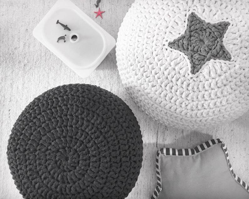 May include: Overhead shot of a white, textured, crocheted pouf with a gray star, a dark gray crocheted pouf, a small gray pillow with striped trim, and a white container with toy fish. The items are on a light-colored surface.