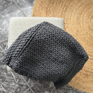 May include: A dark gray, hand-crocheted bag with a textured surface. The bag is sitting on a light gray surface, with a natural fiber rug in the background. The bag's shape is slightly rectangular with a rounded top.