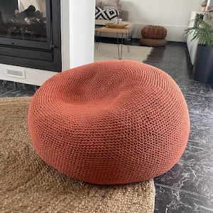 May include: A large, round, terracotta-colored knitted pouf. The pouf is made of thick yarn and sits on a woven jute rug. The pouf is a comfortable seating option for a living room or bedroom. The pouf is approximately 36 inches in diameter.