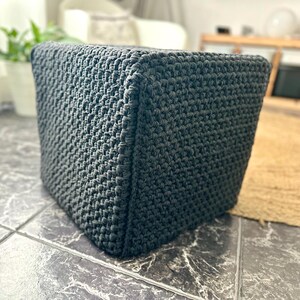 May include: A dark gray, cube-shaped ottoman with a textured, woven appearance. The ottoman is made of a thick, durable material and is suitable for use as a footrest or extra seating. The cube is approximately 16 inches wide and 16 inches tall.