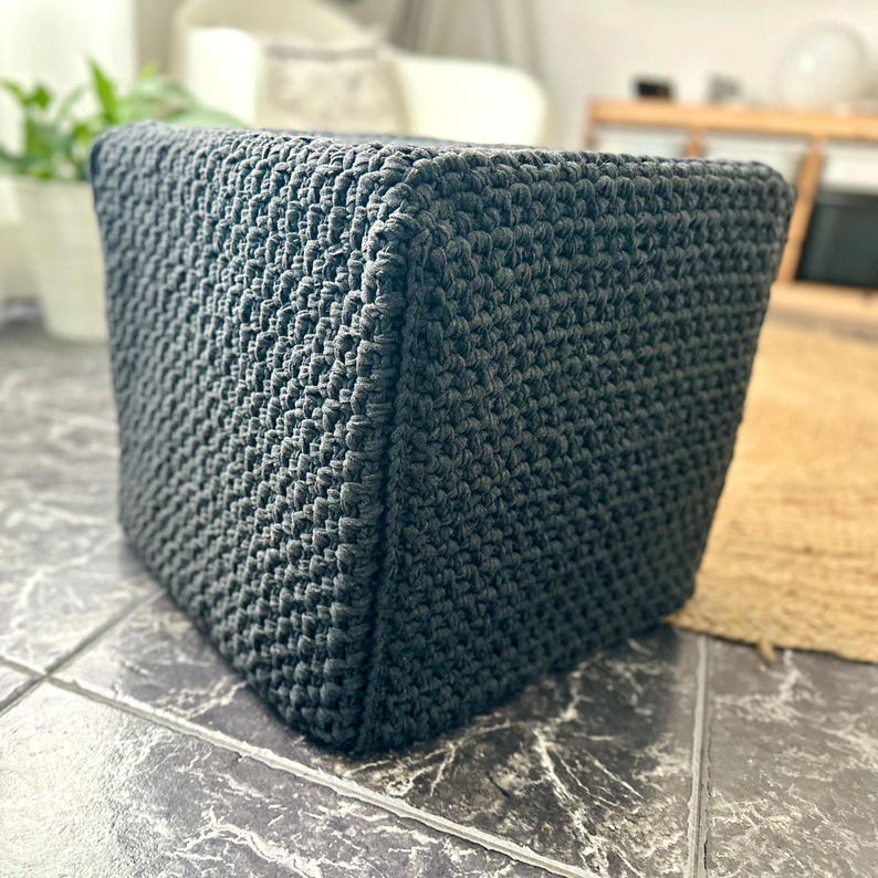 May include: A dark gray, cube-shaped ottoman with a textured, woven appearance. The ottoman is made of a thick, durable material and is suitable for use as a footrest or extra seating. The cube is approximately 16 inches wide and 16 inches tall.