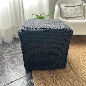 May include: A dark gray, square crochet ottoman. The handmade footstool is made of thick yarn and has a textured surface. The ottoman is placed on a jute rug and a tiled floor.