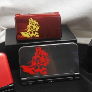 Puede incluir: Una colección de consolas Nintendo 3DS en varios colores, incluyendo rojo, rosa y gris. Cada consola presenta un gráfico estilizado de un guerrero con una espada. Las consolas se exhiben sobre plataformas negras.