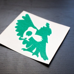 May include: A square white sticker with a green graphic of a bird-like creature. The sticker is on a dark surface. The creature has a large wing and a small head.