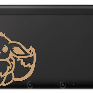 Puede incluir: Consola Nintendo 3DS XL negra con una silueta dorada del personaje Eevee en la tapa superior.