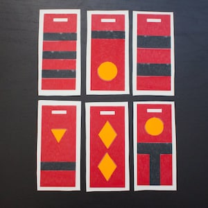 May include: Six rectangular cards with red backgrounds and white borders. Each card features black horizontal stripes or geometric shapes like circles, triangles, and diamonds in yellow. The cards are arranged on a black surface.