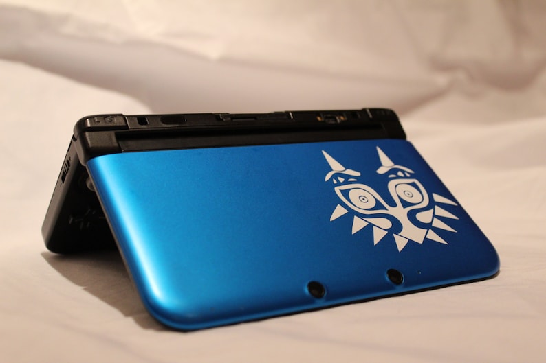 May include: A blue Nintendo 3DS handheld gaming console with a white Majora's Mask design on the top cover.