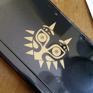May include: A black Nintendo 3DS handheld gaming console with a gold Majora's Mask decal on the top. The console has two cameras on the top.