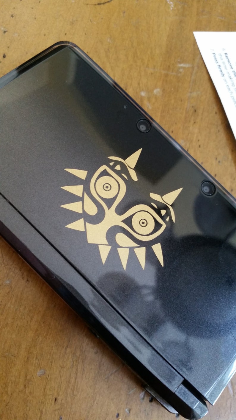 May include: A black Nintendo 3DS handheld gaming console with a gold Majora's Mask decal on the top. The console has two cameras on the top.