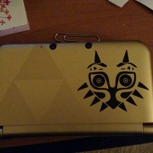 May include: A gold Nintendo 3DS handheld gaming console with a black Majora's Mask decal on the top cover.