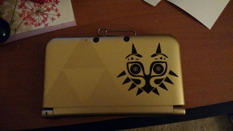 May include: A gold Nintendo 3DS handheld gaming console with a black Majora's Mask decal on the top cover.