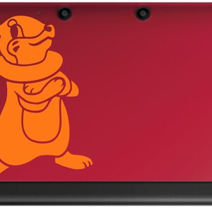 以下が含まれることがあります： 赤いニンテンドー3DS携帯ゲーム機。上部のカバーにオレンジ色の漫画のキャラクターのステッカーが貼られています。キャラクターは、尻尾が巻かれたキツネのような生き物で、友好的な表情をしています。