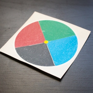 Pode incluir: Um cartão quadrado branco com um design circular colorido. O círculo é dividido em quatro seções: vermelho, verde, azul e cinza. Um pequeno círculo amarelo está no centro. O cartão está sobre uma superfície escura.