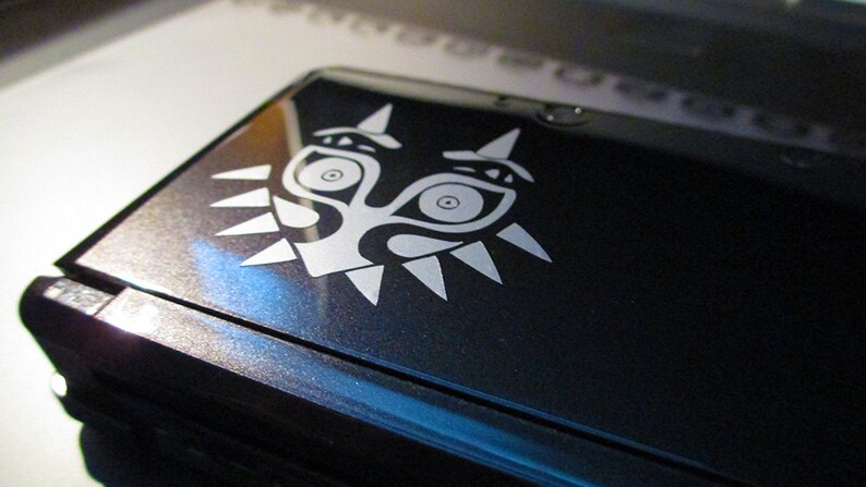 May include: A black Nintendo 3DS handheld gaming console with a white sticker of the Majora's Mask from the Legend of Zelda video game series.