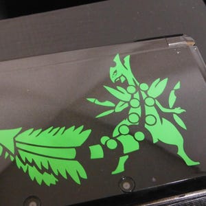 Puede incluir: Consola de juegos portátil negra con un gráfico verde de una criatura estilizada. El diseño presenta una ilustración detallada de una criatura con múltiples extremidades y una larga cola puntiaguda. La consola tiene un acabado brillante.