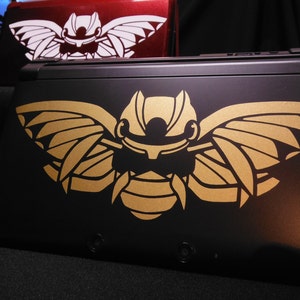 Pode incluir: Um console Nintendo 3DS XL preto com um adesivo metálico dourado de um inseto estilizado com asas. O inseto tem um design detalhado com linhas e padrões intrincados.