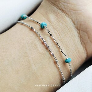 Puede incluir: Un brazalete de cadena plateada con cuentas turquesa. El brazalete está en capas y tiene un diseño delicado. El texto "HENJU BY GRACY" es visible en la parte inferior de la imagen.