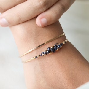 Könnte beinhalten: Zwei goldene Armbänder an einem Handgelenk. Das obere Armband ist ein einfaches Band. Das untere Armband hat einen Cluster aus dunkelblauen Edelsteinen.