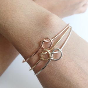 Può includere: Tre braccialetti rigidi con nodo d'oro impilati su un polso. I braccialetti sono in diverse tonalità di oro, tra cui oro rosa, oro giallo e argento.