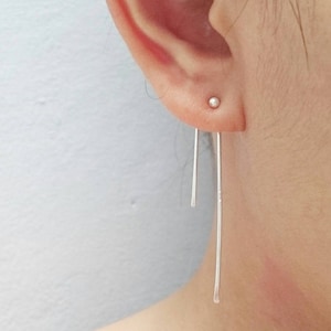 Puede incluir: Un par de pendientes largos y finos de plata con una pequeña bola de plata en la parte superior. Los pendientes cuelgan del lóbulo de la oreja y son muy largos.