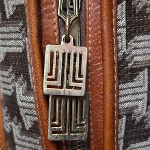 Puede incluir: Primer plano de un bolso marrón y beige con una cremallera dorada y un tirador decorativo dorado. El bolso presenta un patrón geométrico y ribetes de cuero marrón. El tirador de la cremallera tiene un diseño estilizado único.