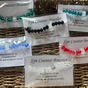May include: Several Syn Counter Bracelets in a range of colours: green, black, blue, red, and clear. Each bracelet has faceted beads, a star charm, and text on the packaging. The text reads "Every crystal is 1/2 Syn, 30 crystals in total, Move the clasp too keep you on track."
