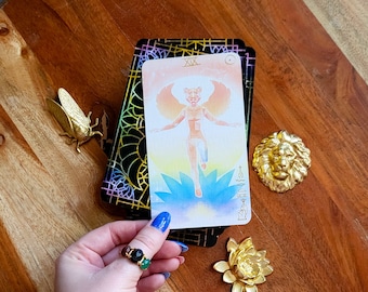 May include: A tarot card with a figure and wings, a deck of cards, and gold-colored decorative items including a lion head, a cicada, and a lotus flower. A hand with a ring and bracelet is holding the card.