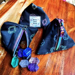 May include: Three black triangular pouches with a gemstone pattern and rainbow zippers. One pouch is open, revealing colorful gemstone dice. The pouches have a label that reads "Glorious Knick Knacks".