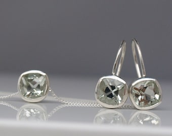 May include: A set of silver earrings and a necklace with light green gemstone pendants. The earrings have a hook closure and the necklace has a chain style.