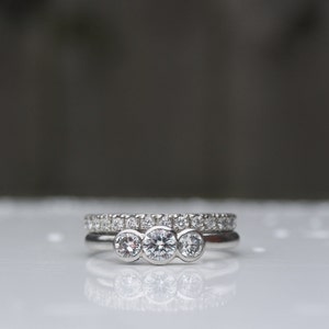 Pode incluir: Um anel de prata com tr&ecirc;s diamantes redondos engastados em fila. O anel est&aacute; empilhado em cima de uma banda de prata com pequenos diamantes.