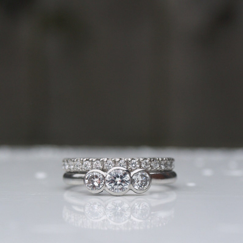Pode incluir: Um anel de prata com tr&ecirc;s diamantes redondos engastados em fila. O anel est&aacute; empilhado em cima de uma banda de prata com pequenos diamantes.