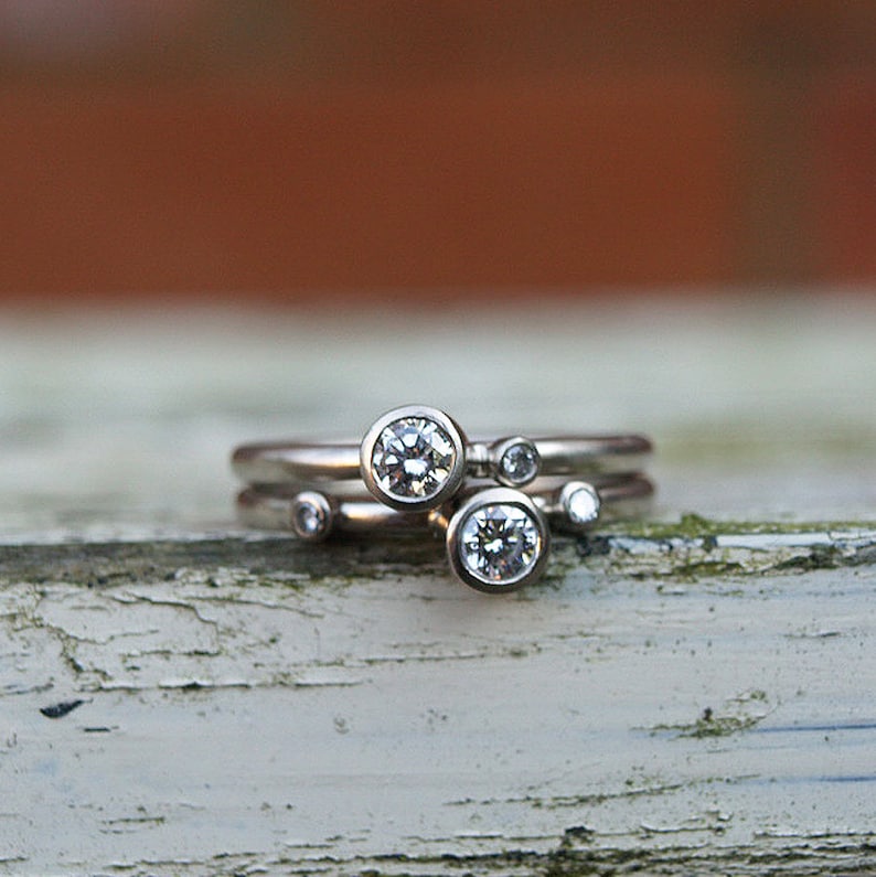 May include: A set of three silver rings with bezel-set diamonds. The largest diamond is in the center, with two smaller diamonds on either side.