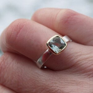 Puede incluir: Un anillo de plata con un engaste de oro y una piedra preciosa azul claro. El anillo est&aacute; en un dedo.