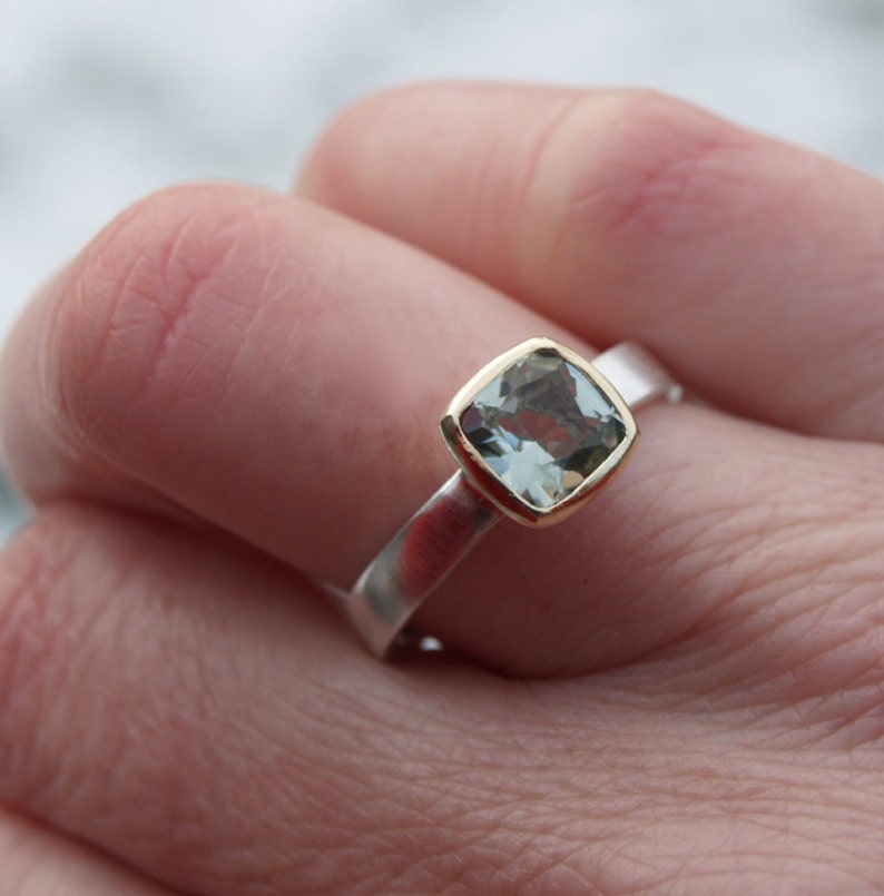 Puede incluir: Un anillo de plata con un engaste de oro y una piedra preciosa azul claro. El anillo est&aacute; en un dedo.