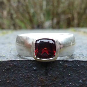 May include: A silver ring with a red gemstone set in a square bezel. The ring has a wide band with a brushed finish.