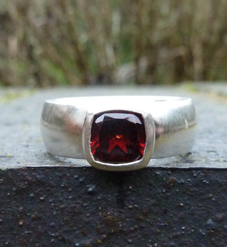 May include: A silver ring with a red gemstone set in a square bezel. The ring has a wide band with a brushed finish.