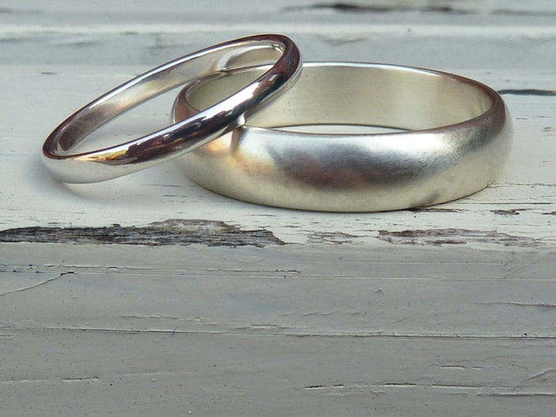Puede incluir: Dos alianzas de boda de plata, una m&aacute;s ancha que la otra, est&aacute;n apiladas una encima de la otra sobre una superficie de madera blanca.