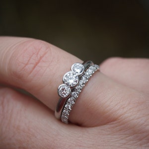 Pode incluir: Um anel de prata com tr&ecirc;s diamantes redondos engastados em fila. O anel &eacute; usado em um dedo com uma segunda banda de prata com pequenos diamantes.