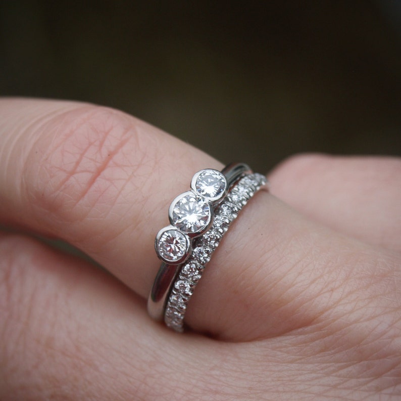 Pode incluir: Um anel de prata com tr&ecirc;s diamantes redondos engastados em fila. O anel &eacute; usado em um dedo com uma segunda banda de prata com pequenos diamantes.
