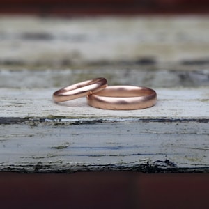 May include: Two gold wedding bands, one slightly larger than the other, are placed side-by-side on a weathered white wooden surface.