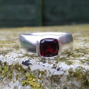 May include: A silver ring with a square-shaped red gemstone set in a bezel setting.