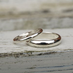 May include: Two silver wedding bands, one slightly wider than the other, are stacked on top of each other on a white wooden surface.