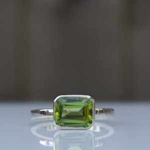 May include: A silver ring with a green gemstone. The gemstone is a square emerald cut.