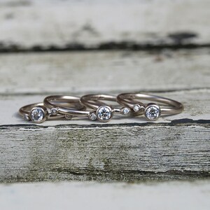 May include: Four silver rings with small round diamonds set in each band. The rings are stacked on top of each other on a weathered wooden surface.