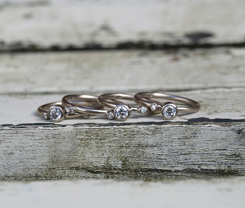 May include: Four silver rings with small round diamonds set in each band. The rings are stacked on top of each other on a weathered wooden surface.