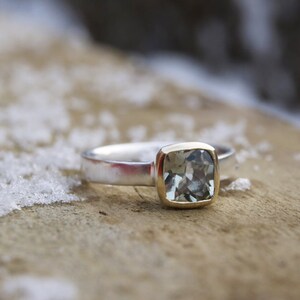 Puede incluir: Un anillo de plata con un engaste de oro y una piedra preciosa azul claro. El anillo est&aacute; sobre una superficie de madera.
