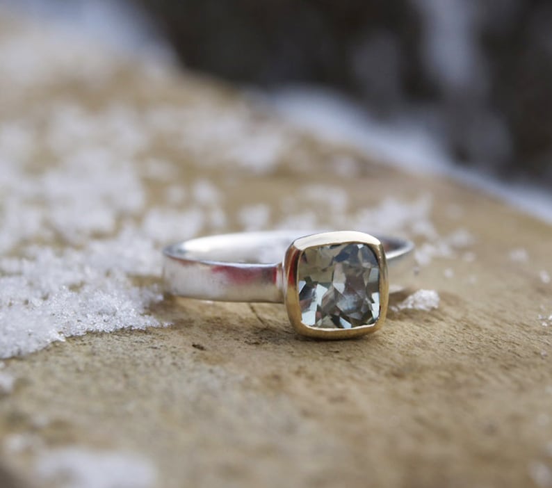 Puede incluir: Un anillo de plata con un engaste de oro y una piedra preciosa azul claro. El anillo est&aacute; sobre una superficie de madera.