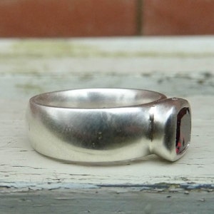 May include: A silver ring with a dark red gemstone set in the band. The ring has a smooth, rounded surface.