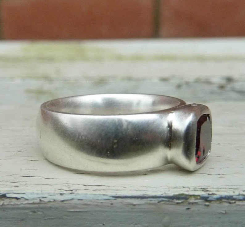 May include: A silver ring with a dark red gemstone set in the band. The ring has a smooth, rounded surface.