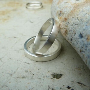 May include: Two silver rings, one stacked on top of the other, with a brushed finish. The rings are on a light gray surface.